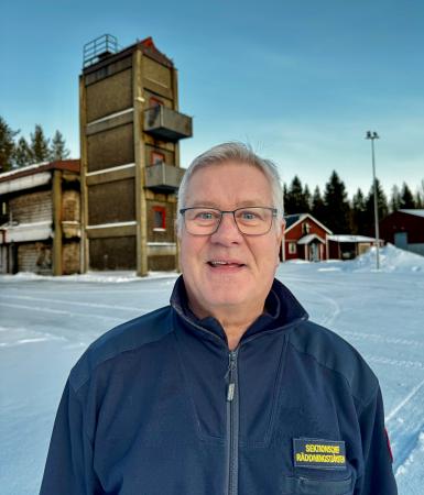 Lars har grått hår och bär förutom glasögon en blå tröja. Han står framför ett brandövningshus som liknar ett torn.