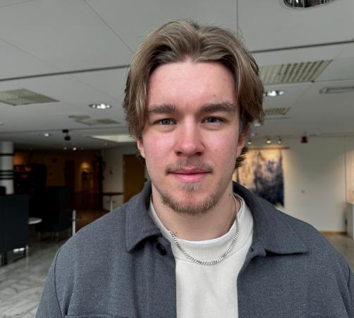 Hugo har en vit tröja och grå skjorta. Han har lite åt hockeyfrilla och getskägg. Han har blå ögon.